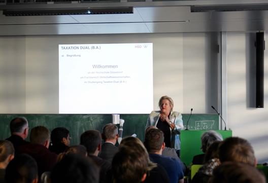 Prof. Dr. Brigitte Grass, die Präsidentin der HSD steht am Kopfende eines Seminarraums hinter einem Pult und begrüßt die Erstsemester.