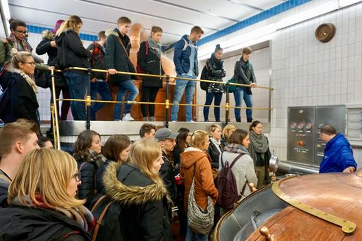 Eine Gruppe Studierender steht in einem gefliesten Raum vor einem großen Kupferkessel. Ein in Blau gekleideter Mitarbeiter der Brauerei erklärt das Brauverfahren.