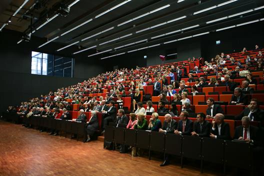 Wir sehen den Audimax der Hochschule Düsseldorf. Auf den aufsteigenden Stuhlreihen sitzen viele festlich gekleidete Menschen unterschiedlichen Alters. Es sind zumeist Absolventen mit ihren Eltern sowie Professoren.
