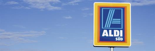 Man sieht ein Hinweisschild für einen Markt der Discounterkette ALDI Süd vor einem blauen Himmel. Das Schild besteht aus einem stilisierten blauen "A" und der weißen Unterschrift "ALDI SÜD". Umrahmt wird das Logo von den Farben rot, orange und gelb.