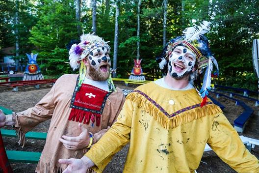 Man sieht zwei junge, als Indianer verkleidete Männer, die gute Laune verbreiten. Im Hintergrund sieht man das Gelände des Camp America, ein Sommercamp für Kinder.