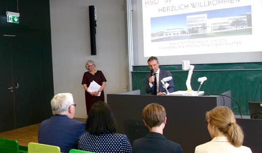 Man sieht die Dekanin des Fachbereiches Wirtschaftswissenschaften, Prof. Dr. Albers und Thomas Geisel, Oberbürgermeister der Stadt Düsseldorf. Sie stehen an der Kopfseite eines Hörsaales. Herr Geisel steht am Rednerpult, Frau Albers links daneben.