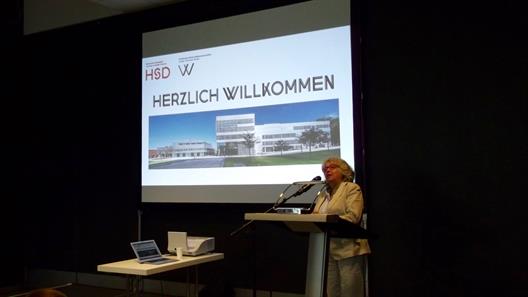 Die Dekanin Prof. Dr. Felicitas Albers steht am Kopfende eines Saales an einem Rednerpult. Im Hintergrund wird eine Präsentation auf einen Hintergrund projiziert. Das aktuelle Chart zeigt die Logos der Hochschule und des Fachbereiches, den Schriftzug "Herzlich willkommen" und eine Außenansicht des Gebäudes, in dem der Fachbereich beheimatet ist.