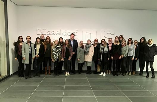Auf dem Bild ist eine Gruppe von 19 jungen Frauen zu sehen, die sich vor einer Wand gruppiert haben. Im Hintergrund befindet sich ein sehr großes plastisches weißes Logo mit dem Schriftzug "Düsseldorf Airport DUS". Ebenfalls auf dem Bild sind Prof. Dr. Kalka und Herr Thomas Kötter.