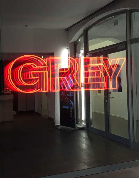 Man sieht eine große Glasscheibe im Eingangsbereich eines Gebäudes. Auf der Scheibe sieht man den Schriftzug "GREY" in roter Neonschrift.